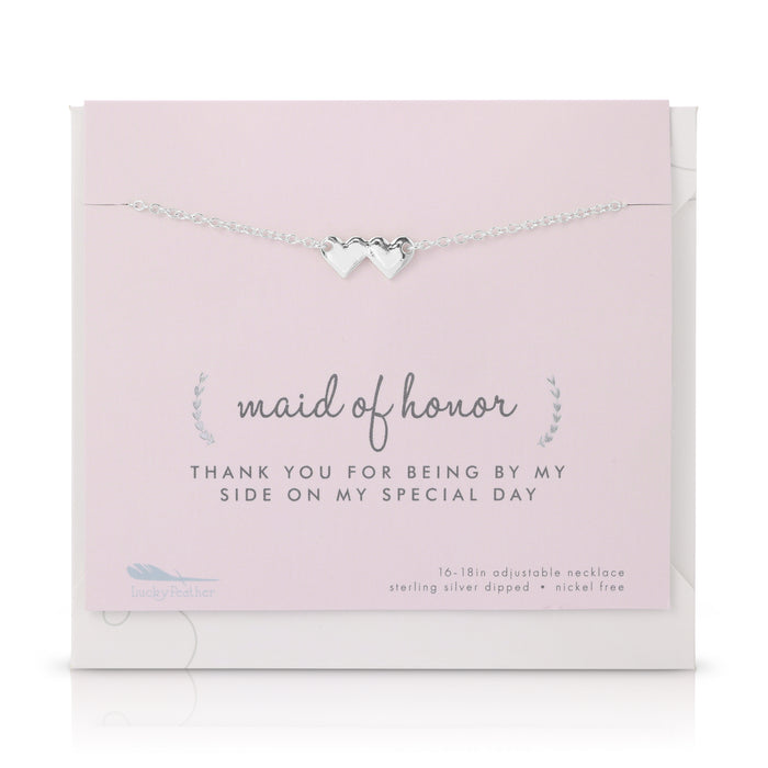 A silver necklace with two hearts is displayed on a pink card with 'maid of honor' printed on it.