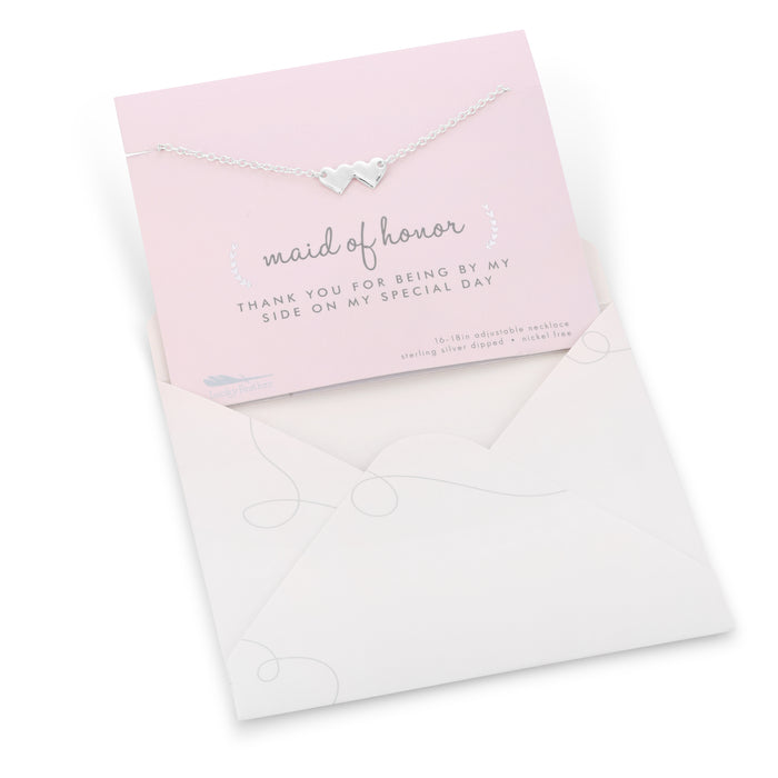 A silver necklace with two heart pendants is displayed on a 'maid of honor' thank you card inside a light-colored envelope.