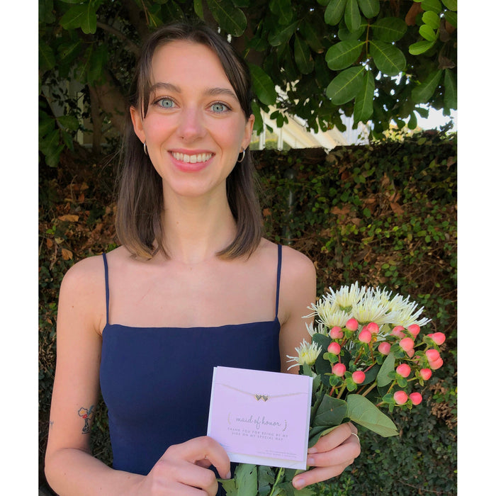 A smiling woman in a navy dress holds a bouquet of white and pink flowers and a 'Maid of Honor' thank you card.