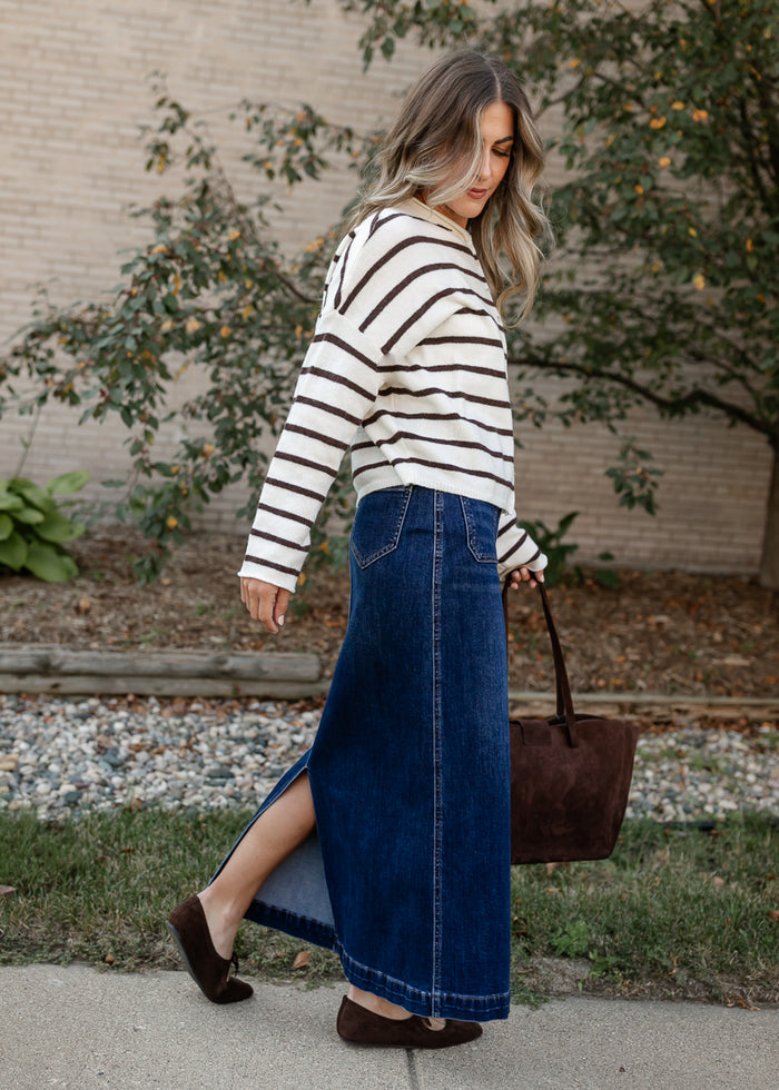 A woman wearing a cream and brown striped knit long-sleeve top and a high-waisted blue denim maxi skirt with a side slit, walking outdoors while carrying a brown leather tote bag.