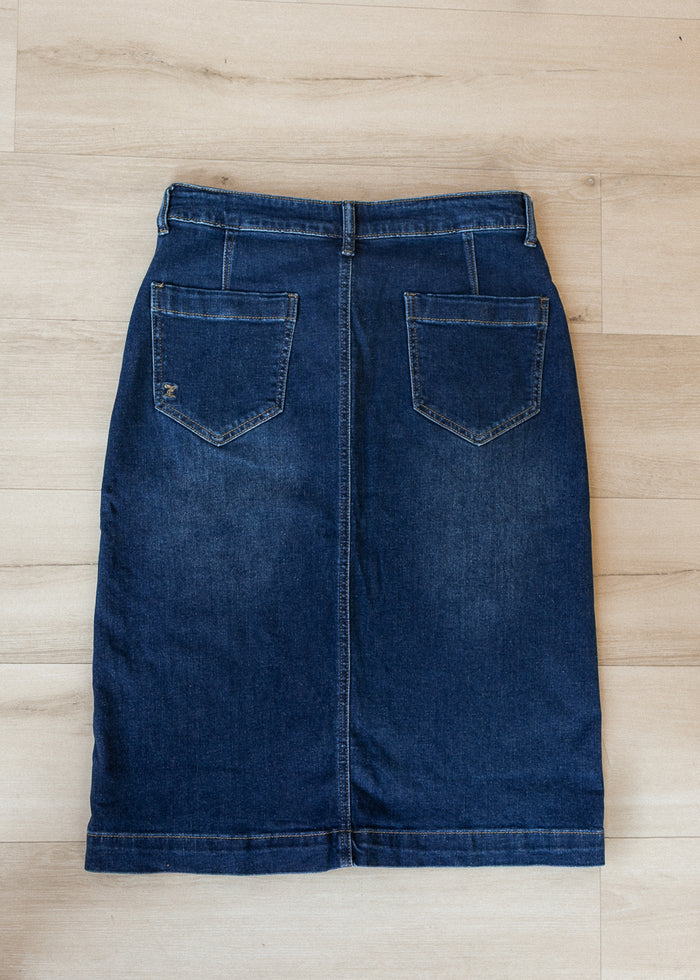 A dark blue denim skirt with two back patch pockets, laid flat on a light wood plank floor.