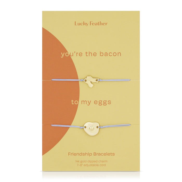 Two 'Lucky Feather' friendship bracelets with gold-dipped bacon and egg charms on adjustable cords, presented on a decorative card with the text 'you're the bacon to my eggs'.