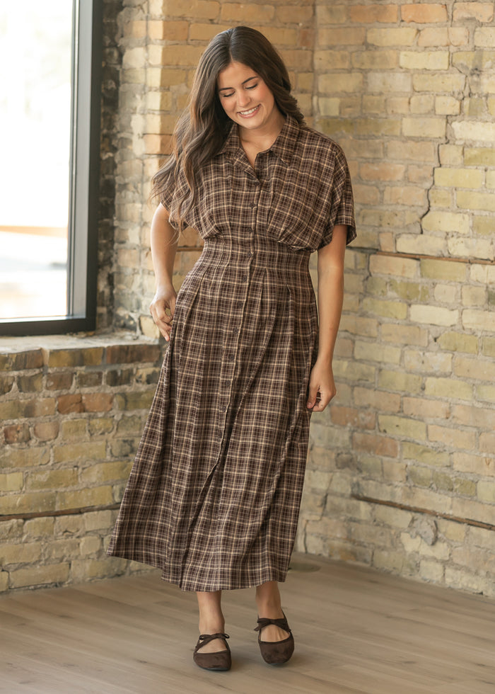 A woman wearing a brown and beige plaid midi shirt dress with short sleeves, standing in a room with a brick wall and wood floor.