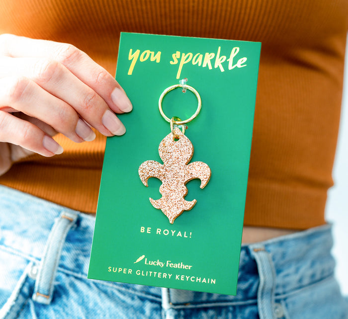A person holds a 'Lucky Feather' super glittery keychain shaped like a fleur-de-lis against an orange shirt and blue jeans.