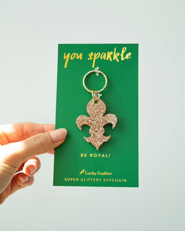 A hand holds a green card featuring a fleur-de-lis keychain with glitter.