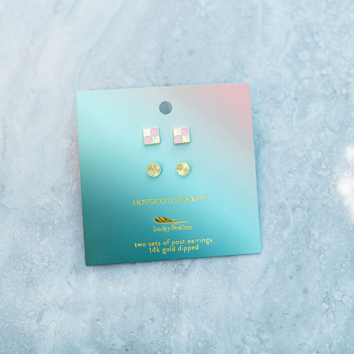 Two sets of post earrings, one pink and white checkered and the other gold swirl, are displayed on a turquoise and pink card with the logo 'Lucky Feather.'