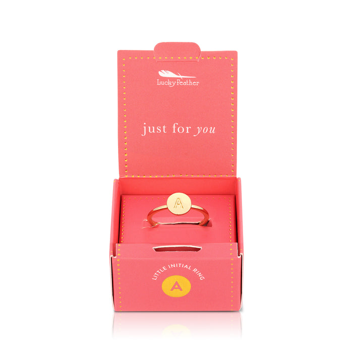 A gold ring with the letter 'A' inside a circular setting sits inside a pink 'Lucky Feather' box.