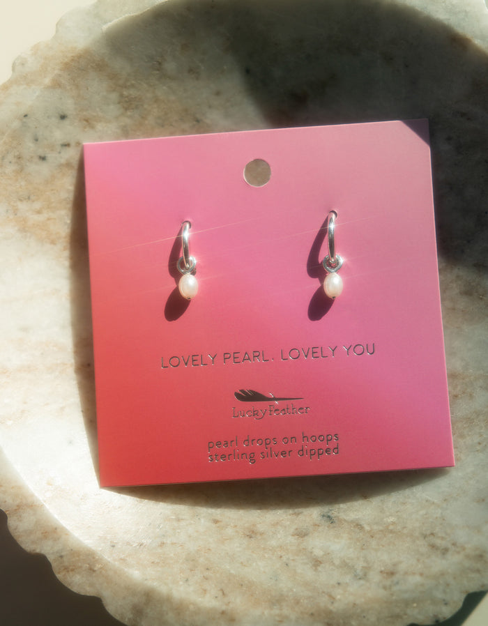 A pair of 'Lucky Feather' pearl drop earrings are displayed on a pink card.