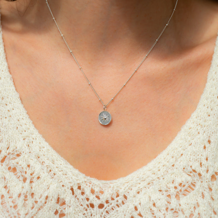 A person wearing a silver chain necklace with a round pendant featuring a sunburst design.