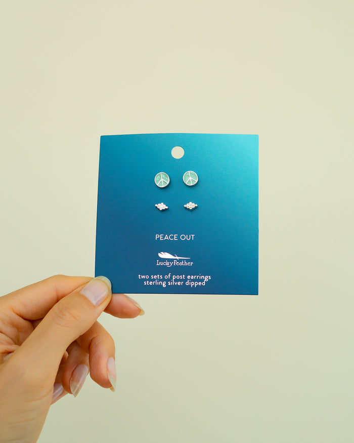 A hand holds a blue card with 'Peace Out' and 'Lucky Feather' printed on it, displaying two sets of post earrings: peace signs and clouds.