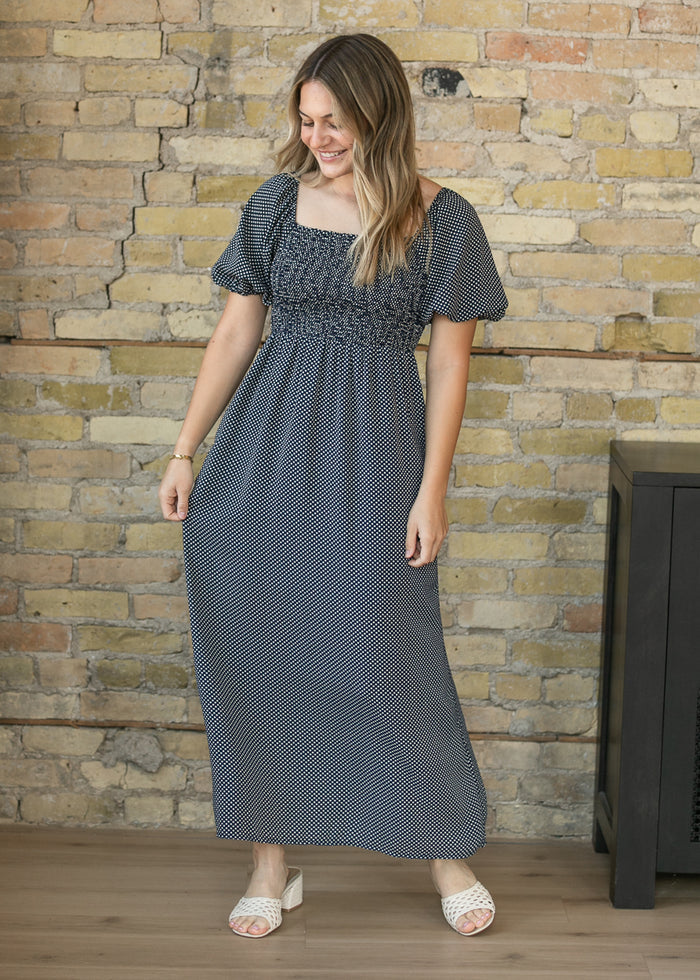 A woman wearing a dark gray and white micro-patterned maxi dress with a smocked bodice and short puff sleeves, standing in front of a rustic brick wall.