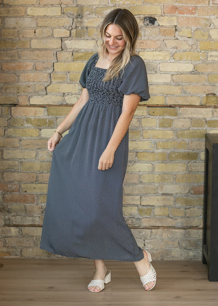 A woman modeling a dark gray and white micro-dot maxi dress with a smocked bodice, standing in front of a rustic brick wall.