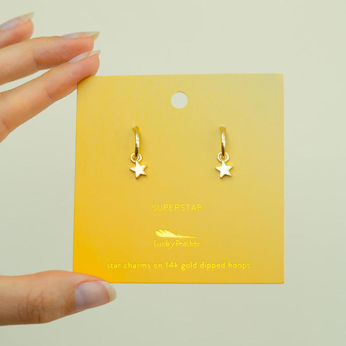 A pair of gold star-shaped charm earrings on a yellow card held by a hand.